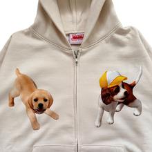 Load image into Gallery viewer, An off-white zip-up hoodie featuring two printed puppy graphics on the chest. On the left chest is a light tan puppy, and on the right chest is a brown and white puppy wearing a yellow and white cap.
